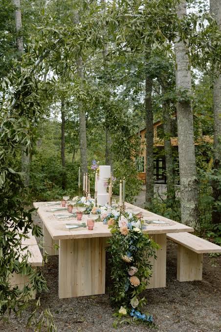 A farm table with a tiered wedding cake and floral garland set among the trees